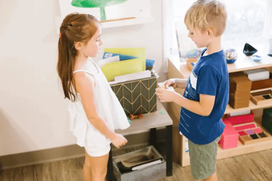 Two Montessori children standing face to face in a classroom, practicing a social interaction lesson with one child offering an item to the other.