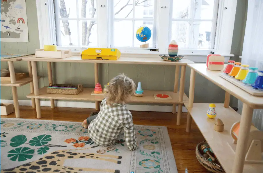 Toddler sitting on a rug independently choosing Montessori materials from low wooden shelves in a calm, light-filled classroom.