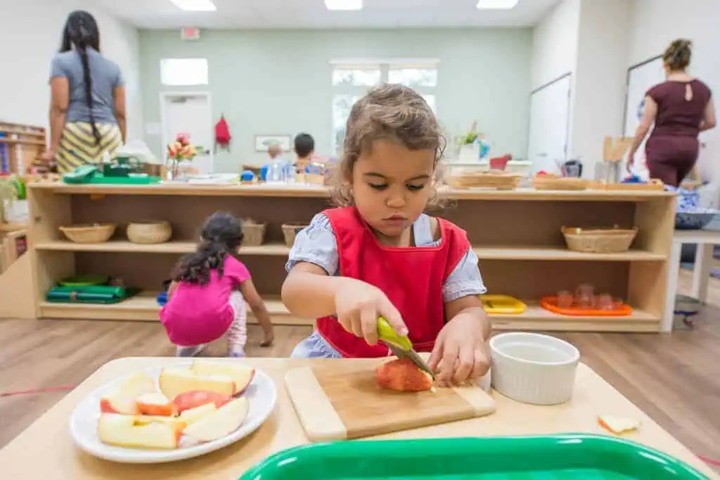 Toddler slicing fruit independently in a Montessori classroom, engaging in real food preparation instead of completing a chore chart task.
