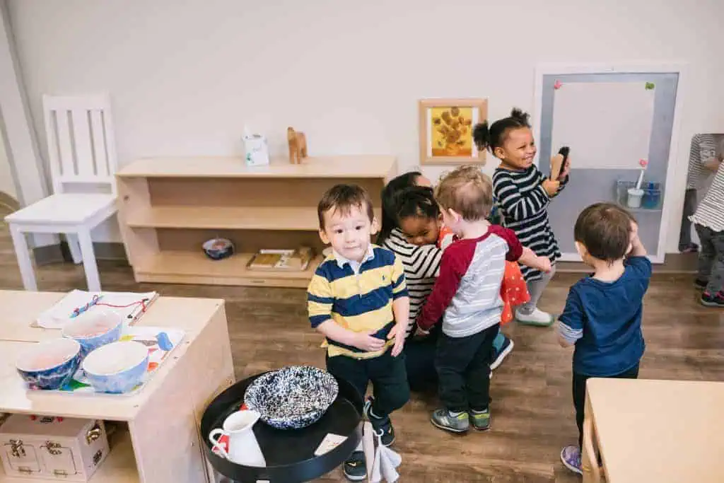 Several toddlers gather in a classroom near a low table with practical life materials, while one child looks toward the camera.