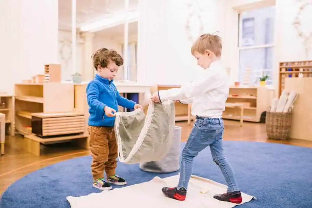 Two young children hold a cloth together on a rug in a Montessori classroom, practicing coordinated movement.