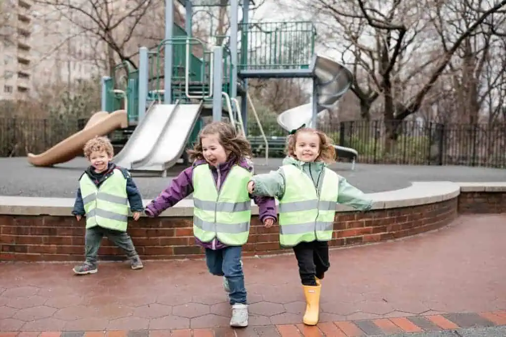 Three young children in bright reflective vests run together near a playground structure on an outdoor play area.