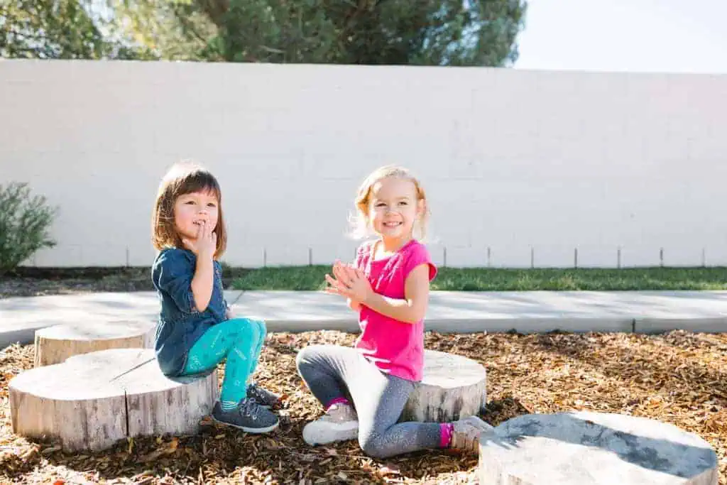 Two young girls sit on wooden stumps outdoors, smiling and clapping in a natural play area.