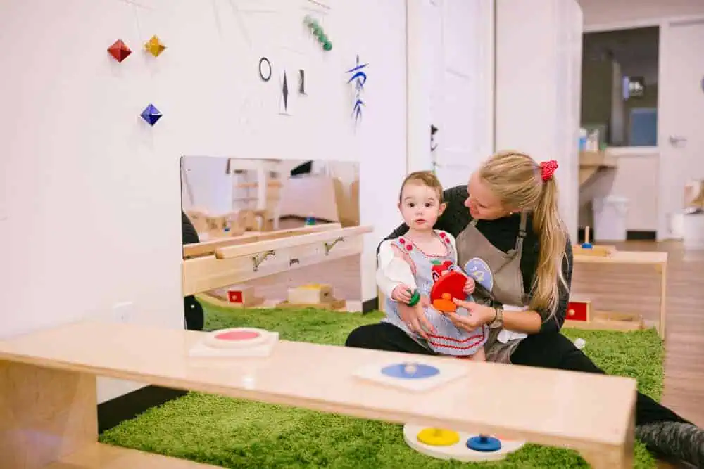 Guidepost Montessori Guide sitting with an infant on a green rug, introducing sensorial materials in a calm classroom environment.