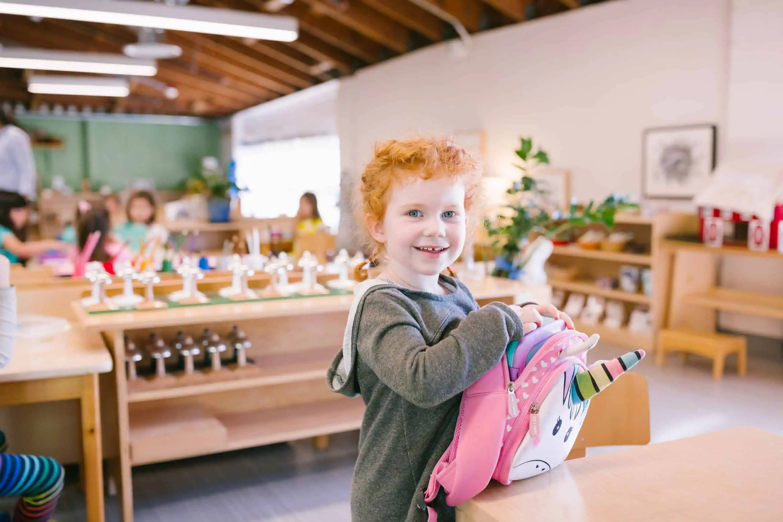 Young girl with curly red hair smiling in a Montessori classroom, holding a pink unicorn backpack