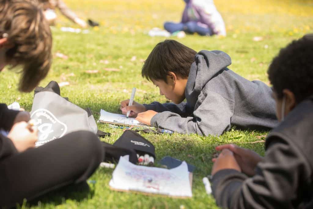 Children studying outdoors on grass, practicing independence in learning activities.
